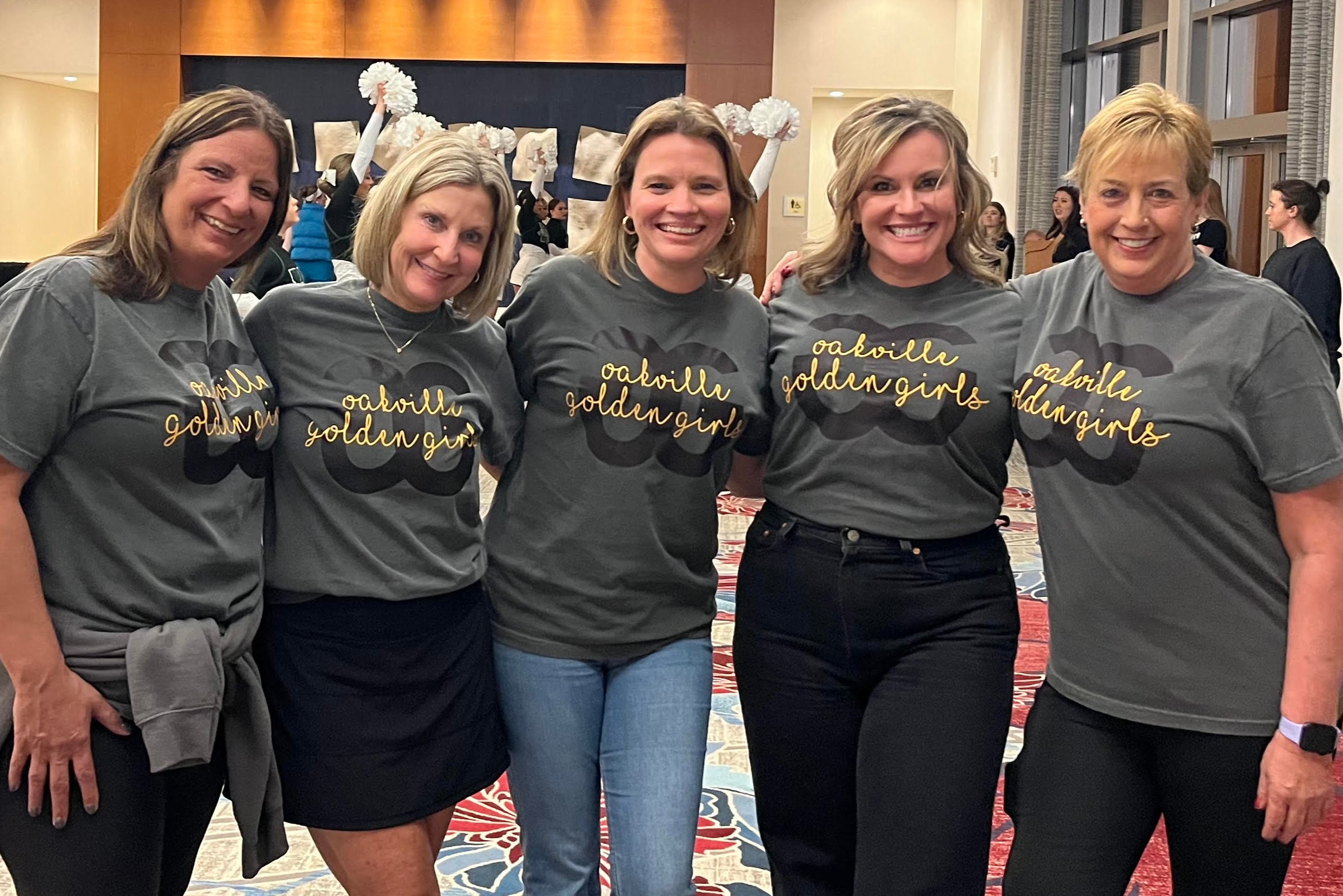 Five women in matching gray t-shirts standing together in a hotel lobby.